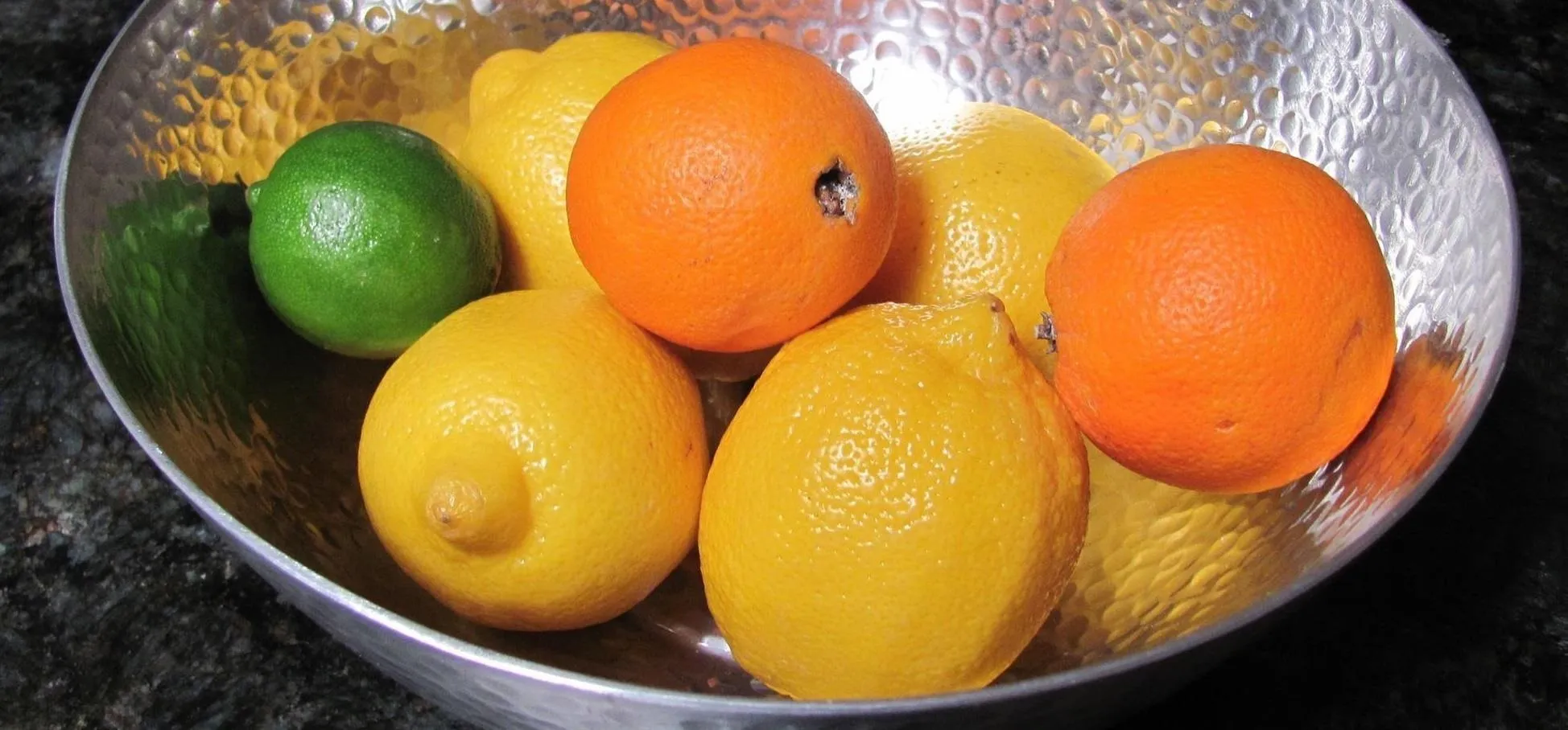 Hammered metal bowl filled with fresh lemons, oranges, and a lime, resting on a dark granite countertop.