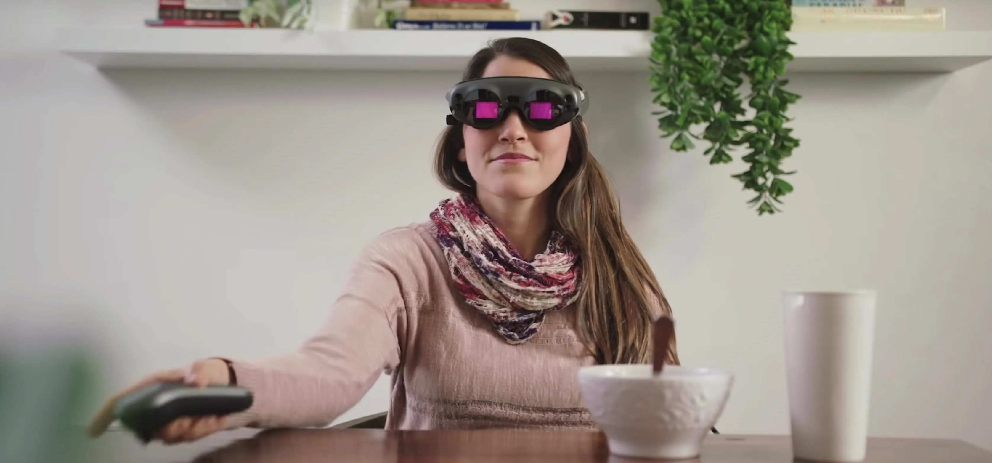 Woman wearing augmented reality glasses, holding a remote control, seated at a table with a bowl.