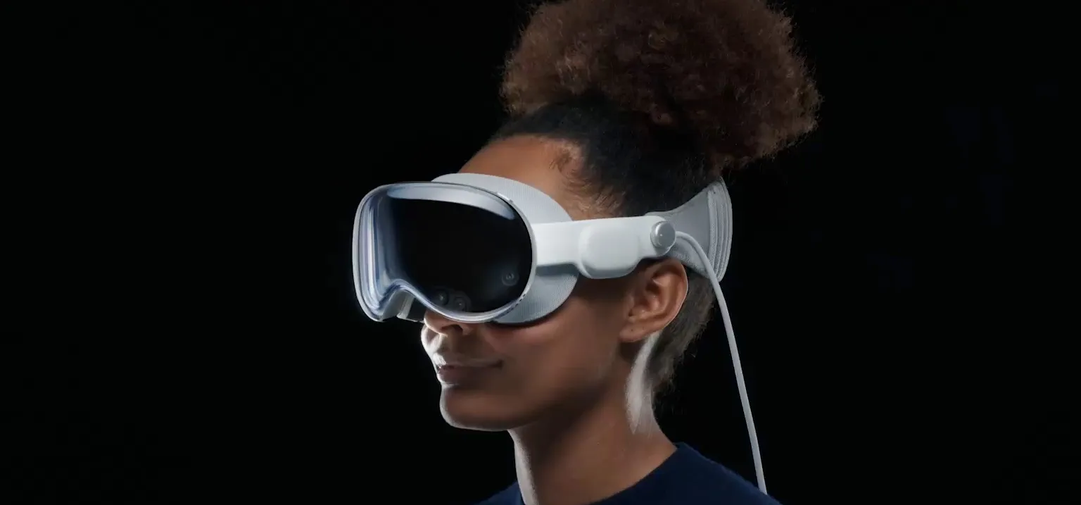 A person wearing Apple’s Vision Pro headset with a tethered cable, shown in a dark studio setting.
